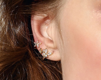 Puede incluir: Primer plano de una oreja con tres pendientes de oro en forma de flor con cristales transparentes. Los pendientes están agrupados en el lóbulo de la oreja.
