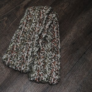 May include: A pair of knitted leg warmers in a brown, white, and green color scheme. The leg warmers are folded in half and have a textured knit pattern.