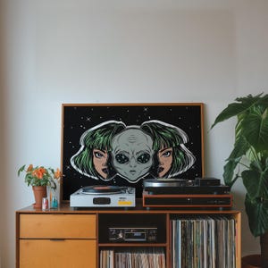 May include: A framed art print featuring an alien head with two female faces on a black background with stars. The artwork is displayed on a wooden cabinet with two record players, a stereo, and a collection of vinyl records. A potted plant is on the right.