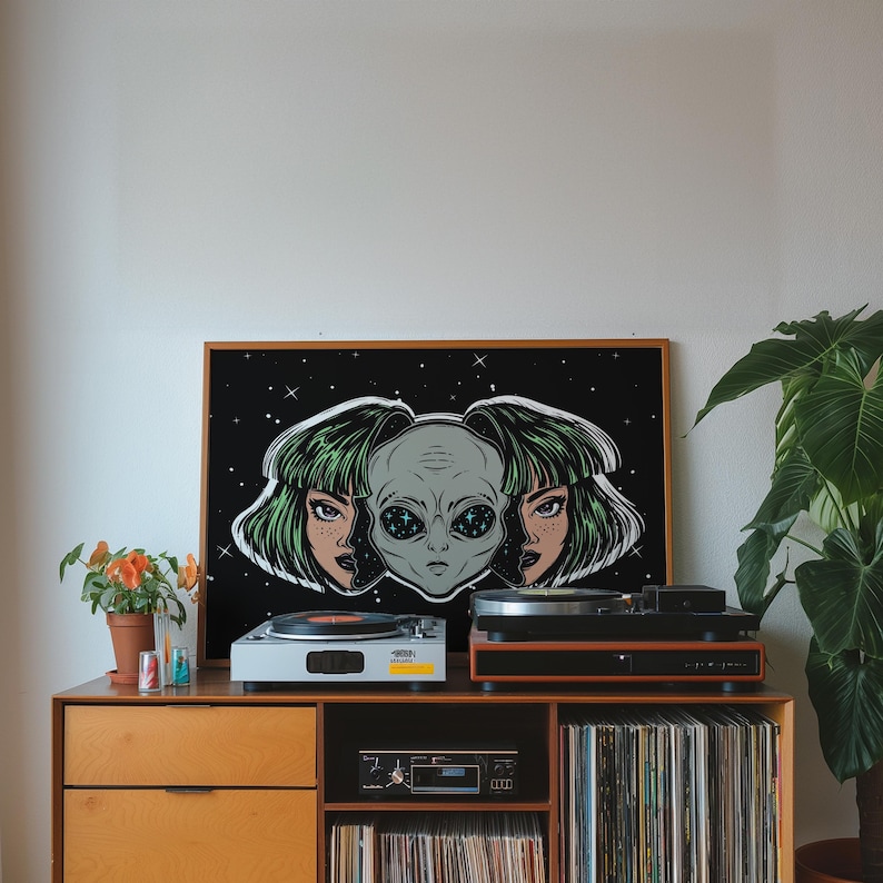 May include: A framed art print featuring an alien head with two female faces on a black background with stars. The artwork is displayed on a wooden cabinet with two record players, a stereo, and a collection of vinyl records. A potted plant is on the right.