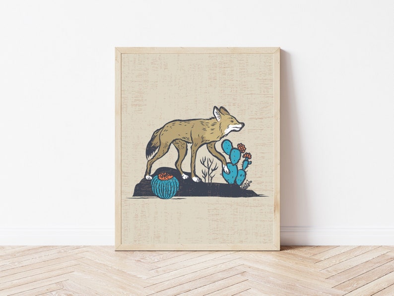 May include: A framed art print featuring a tan coyote walking across a dark rock formation. The scene includes a blue prickly pear cactus with orange flowers and a small round blue cactus with an orange top, all set on a beige background.