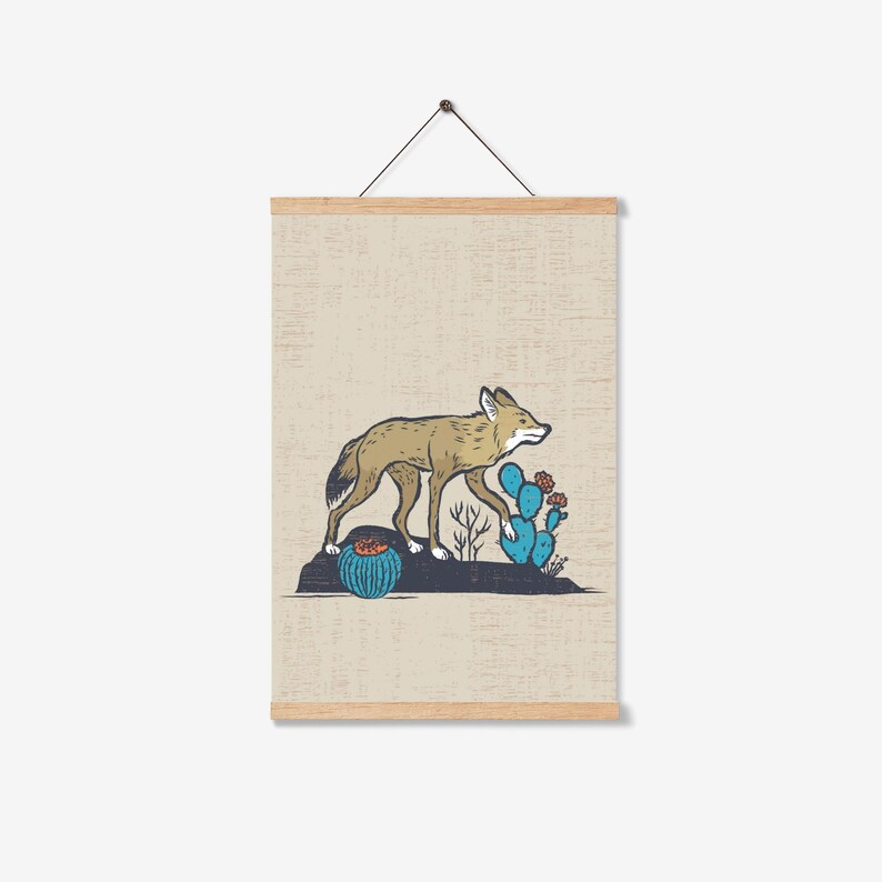 May include: A framed print featuring a coyote walking past cacti. The coyote is tan and brown, the cacti are blue and green, and the background is a light beige. The print is held by wooden magnetic poster hangers.