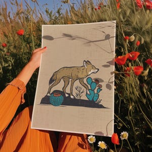 May include: A tan and brown illustration of a coyote walking past a blue cactus and desert plants. The artwork is on a beige background. The image is held in front of a field of red poppies and green grass.