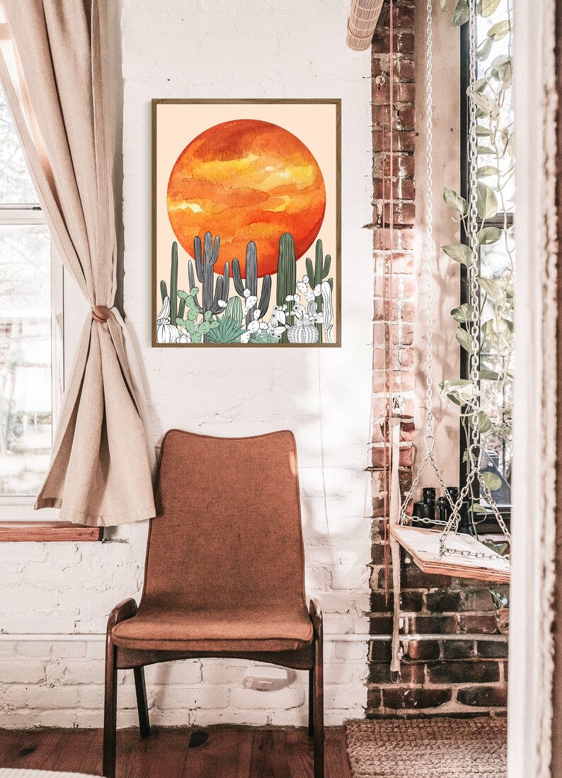 May include: A framed art print featuring a watercolor illustration of a desert landscape. The artwork depicts a large orange sun above various cacti in shades of green and gray. The print is displayed in a room with a chair and a window.
