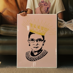May include: A framed art print with a portrait of a woman in glasses and a decorative collar, rendered in black and white. A gold crown with star details and radiating lines sits above her head. The background is a pale pink.