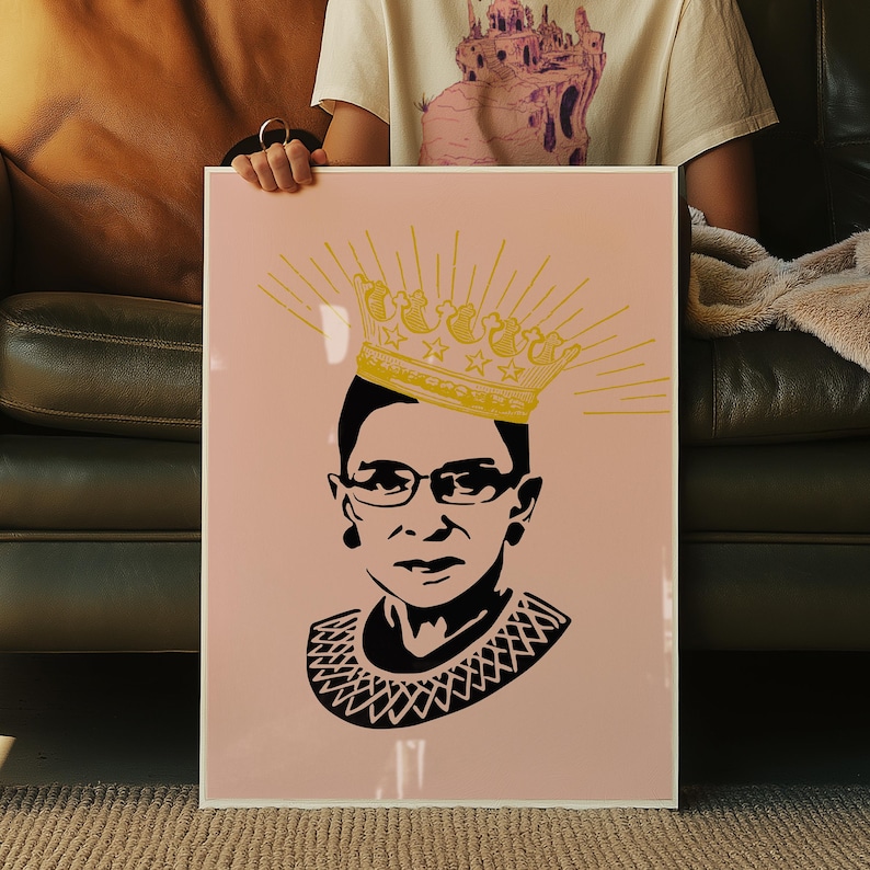May include: A framed art print with a portrait of a woman in glasses and a decorative collar, rendered in black and white. A gold crown with star details and radiating lines sits above her head. The background is a pale pink.