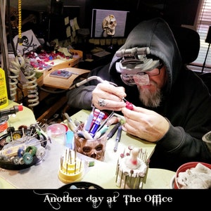 May include: A person wearing a black hooded sweatshirt and glasses works on a red wax sculpture at a workbench. The workbench is covered in tools and supplies, including a skull-shaped container, a coil of wire, and a variety of small tools. The text "Another day at The Office" is visible at the bottom of the image.