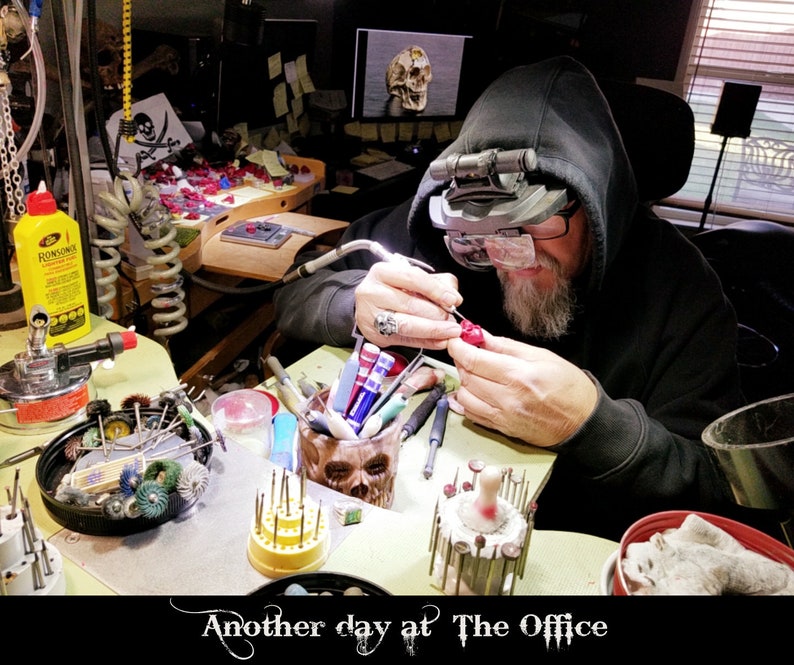 May include: A person wearing a black hooded sweatshirt and glasses works on a red wax sculpture at a workbench. The workbench is covered in tools and supplies, including a skull-shaped container, a coil of wire, and a variety of small tools. The text "Another day at The Office" is visible at the bottom of the image.