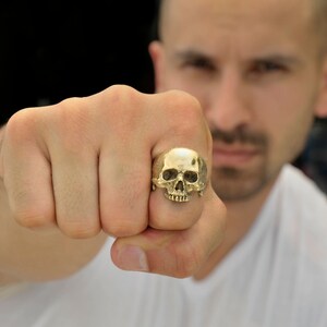 May include: A gold skull ring with a detailed design. The ring is worn on the finger of a person's hand.