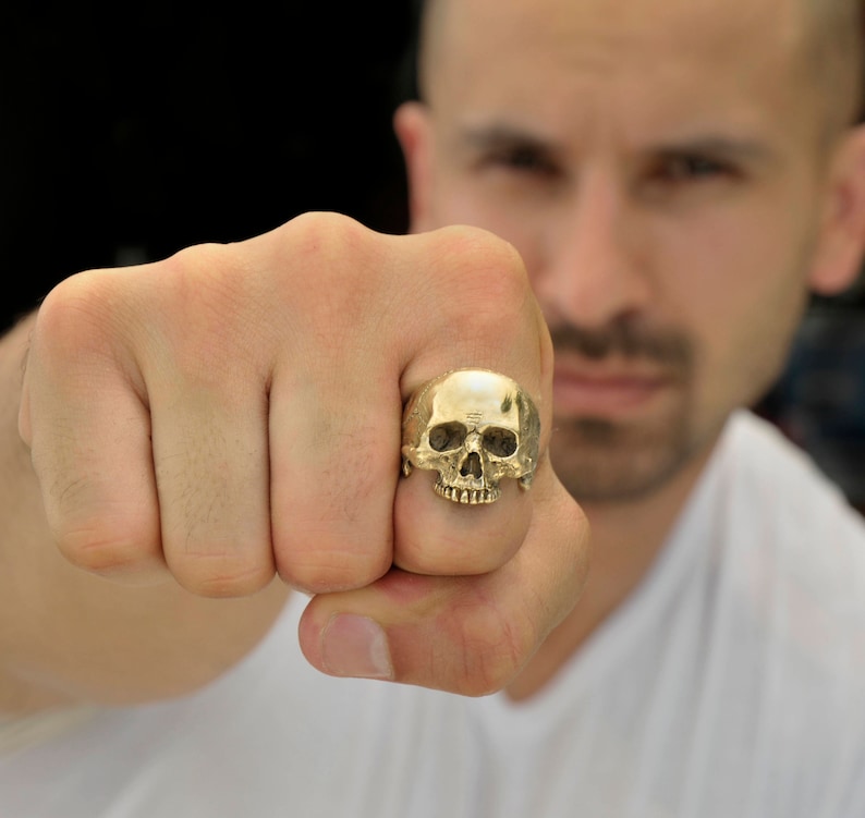 May include: A gold skull ring with a detailed design. The ring is worn on the finger of a person's hand.