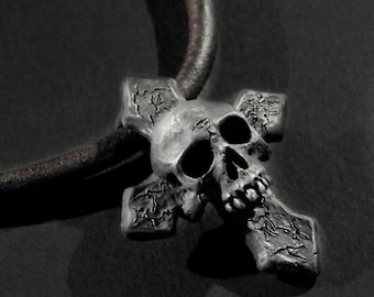 May include: A silver skull pendant with a cross design. The skull has a detailed face with a cracked texture. The pendant is on a brown leather cord.