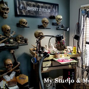 May include: A cluttered workspace with a workbench and tools, including a microscope, a vacuum system, and a variety of jewelry-making tools. The walls are decorated with several skull models, including a large ape skull and a sign that reads "JOHNNY 10 RINGS".