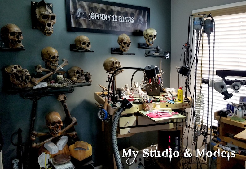 May include: A cluttered workspace with a workbench and tools, including a microscope, a vacuum system, and a variety of jewelry-making tools. The walls are decorated with several skull models, including a large ape skull and a sign that reads "JOHNNY 10 RINGS".