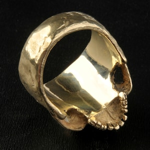 May include: A gold skull ring with a textured band. The ring is designed to look like a human skull with a detailed face.