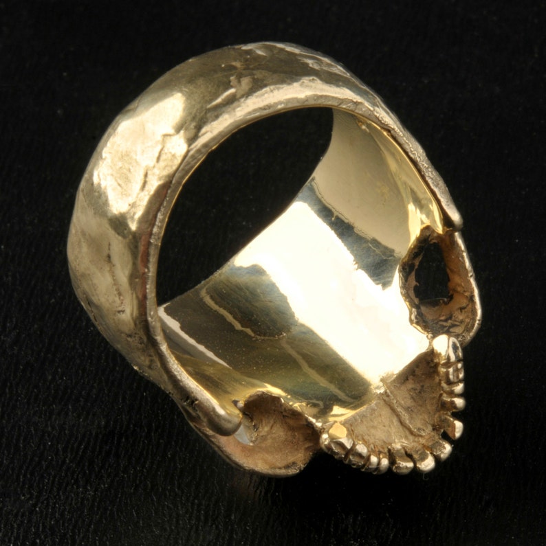 May include: A gold skull ring with a textured band. The ring is designed to look like a human skull with a detailed face.