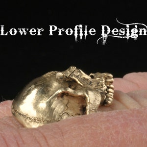 May include: A gold-colored skull ring with a lower profile design. The ring is worn on a finger.