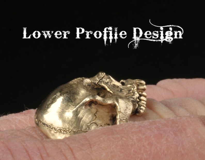 May include: A gold-colored skull ring with a lower profile design. The ring is worn on a finger.