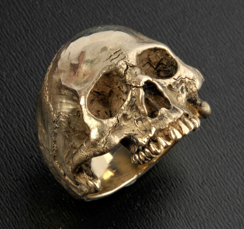 May include: A gold-colored skull ring with a detailed design. The ring is made of metal and has a textured surface.