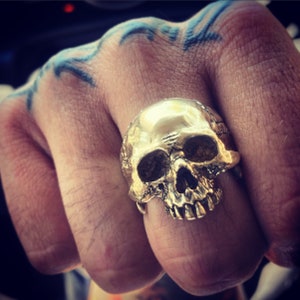 May include: A gold skull ring with detailed features. The ring is worn on a finger with a blue tattoo.