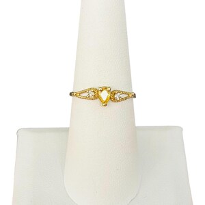 May include: A gold ring with a pear-shaped yellow gemstone at the center, flanked by two smaller clear stones. The band features a delicate, openwork design. The ring is displayed on a white jewelry stand.