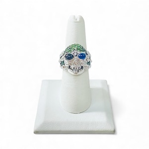 May include: A silver skull ring with green, blue, and clear gemstones. The ring features a skull design with green gems on the forehead, blue and dark blue gems for the eyes, and clear gems around the skull's edges.