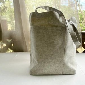May include: A large, light gray canvas tote bag with two long handles. The bag has a large pocket on the front.