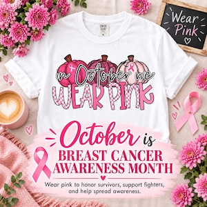 Op de afbeelding: Een wit t-shirt met een afbeelding van drie pompoenen in roze tinten, voorzien van de tekst "In October we wear pink" in een gestileerd lettertype. Het ontwerp bevat een roze lint-symbool op de pompoenen voor de borstkankermaand.