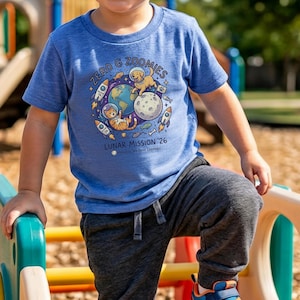 Puede incluir: Una camiseta azul con un gráfico colorido que presenta un gato, un perro, la Tierra, la luna y cohetes. El texto en la camiseta dice "ZERO G ZOOMIES LUNAR MISSION '26". La camiseta la lleva un niño.