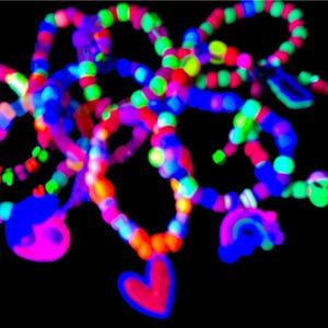 May include: A collection of colorful beaded bracelets and necklaces against a black background. The jewelry features a variety of bright colors, including pink, blue, green, and red. Some pieces have heart and rainbow charms.