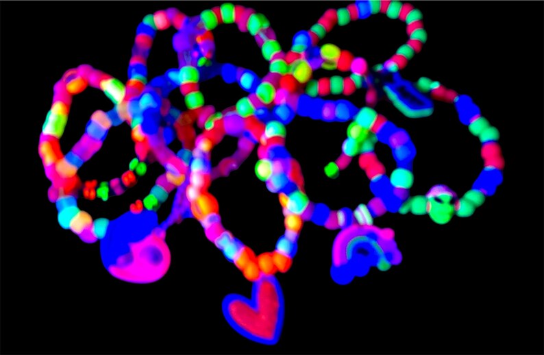 May include: A collection of colorful beaded bracelets and necklaces against a black background. The jewelry features a variety of bright colors, including pink, blue, green, and red. Some pieces have heart and rainbow charms.