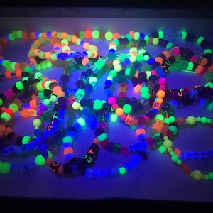 May include: A collection of colorful beaded bracelets, glowing under a black light. The bracelets feature neon green, orange, pink, and blue beads. Some bracelets have letter beads spelling out words like "TRANCE" and "ZOOM".