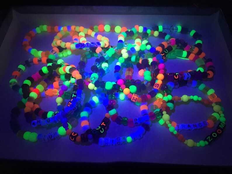 May include: A collection of colorful beaded bracelets, glowing under a black light. The bracelets feature neon green, orange, pink, and blue beads. Some bracelets have letter beads spelling out words like "TRANCE" and "ZOOM".