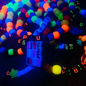 May include: A collection of colorful beaded bracelets and accessories, glowing under a black light. The bracelets feature neon orange, blue, green, and yellow beads. Some bracelets spell out words like "LIVE LIFE HAPPY" and "2025".