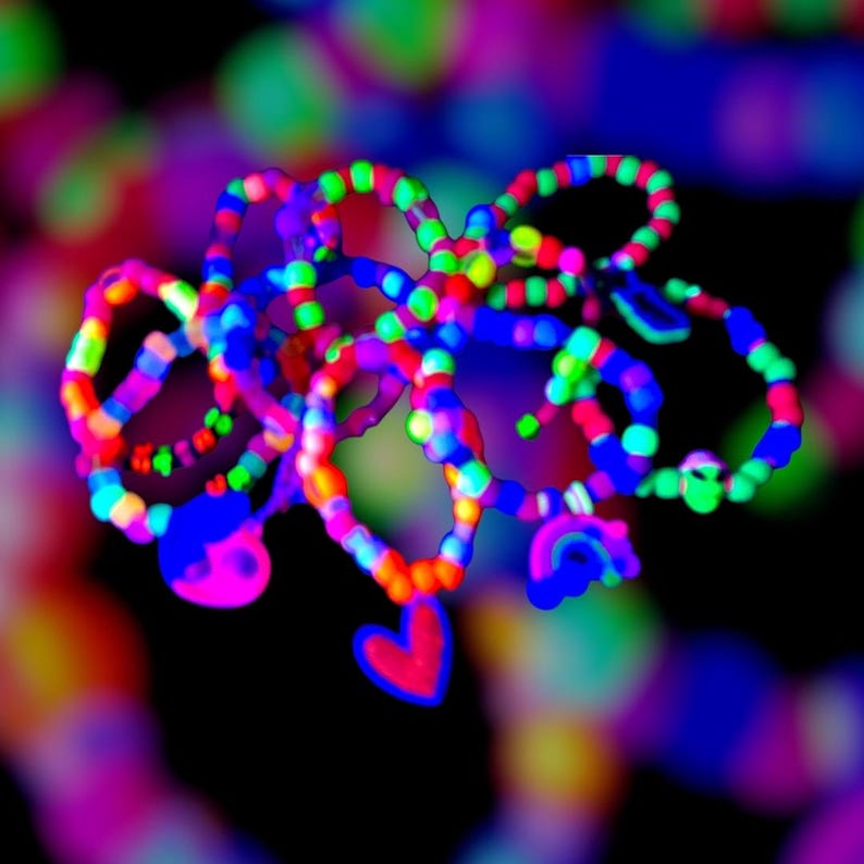 May include: A collection of colorful bracelets with heart and rainbow charms. The bracelets are adorned with vibrant neon colors including red, blue, green, and yellow. The image has a blurred background with bokeh effects.