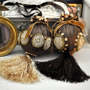 May include: Two brown fabric ornaments with gold and white beaded embellishments and tassels. The ornaments are hanging from a gold frame with a cherub figurine.