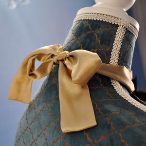 May include: Close-up of a teal-colored garment with a gold satin bow. The fabric has a gold geometric pattern and is trimmed with white lace. The garment is displayed on a white mannequin.