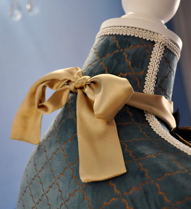 May include: Close-up of a teal-colored garment with a gold satin bow. The fabric has a gold geometric pattern and is trimmed with white lace. The garment is displayed on a white mannequin.