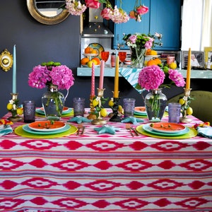 Peut inclure: Une nappe rose et blanche avec un motif g&eacute;om&eacute;trique et un bord festonn&eacute;. La table est dress&eacute;e avec des assiettes, des verres et des chandeliers. Il y a des fleurs roses dans un vase et des citrons sur la table.