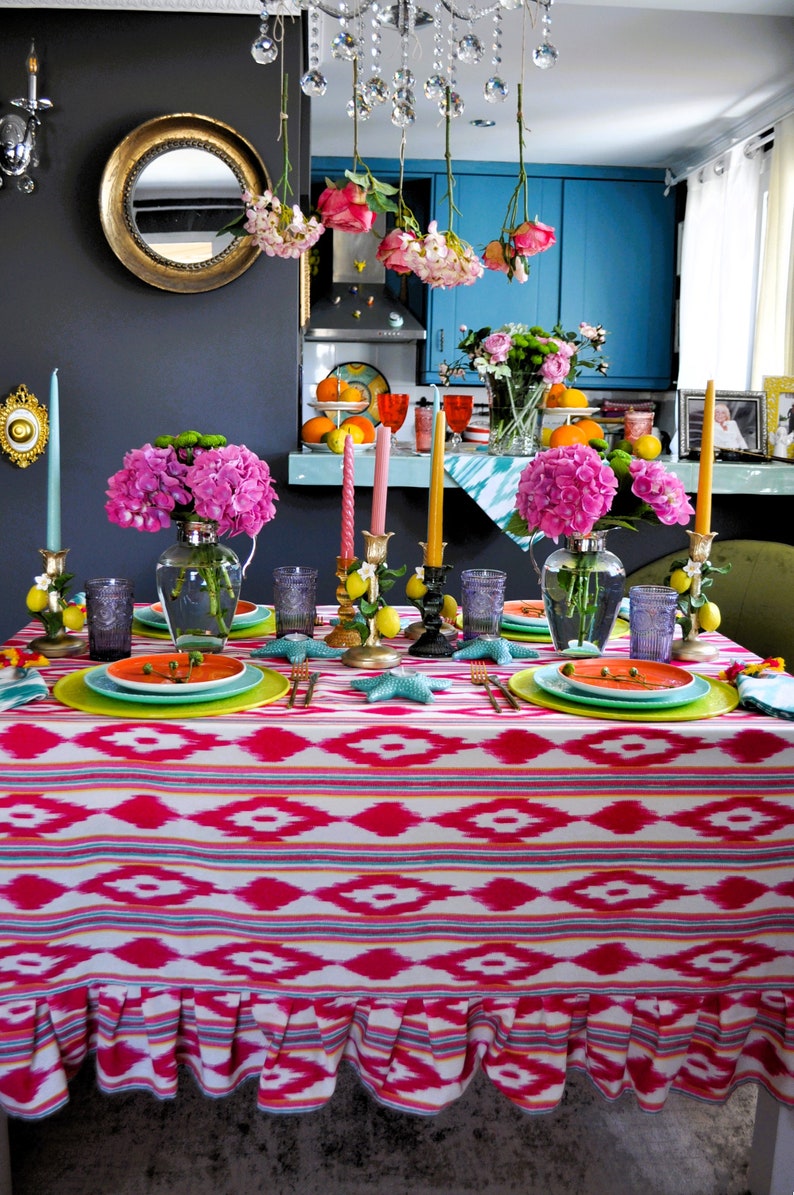 Peut inclure: Une nappe rose et blanche avec un motif g&eacute;om&eacute;trique et un bord festonn&eacute;. La table est dress&eacute;e avec des assiettes, des verres et des chandeliers. Il y a des fleurs roses dans un vase et des citrons sur la table.