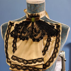 May include: A cream-colored satin ascot with black lace trim and a black lace overlay. The ascot features a brown velvet bow with a green jewel and is displayed on a blue mannequin.