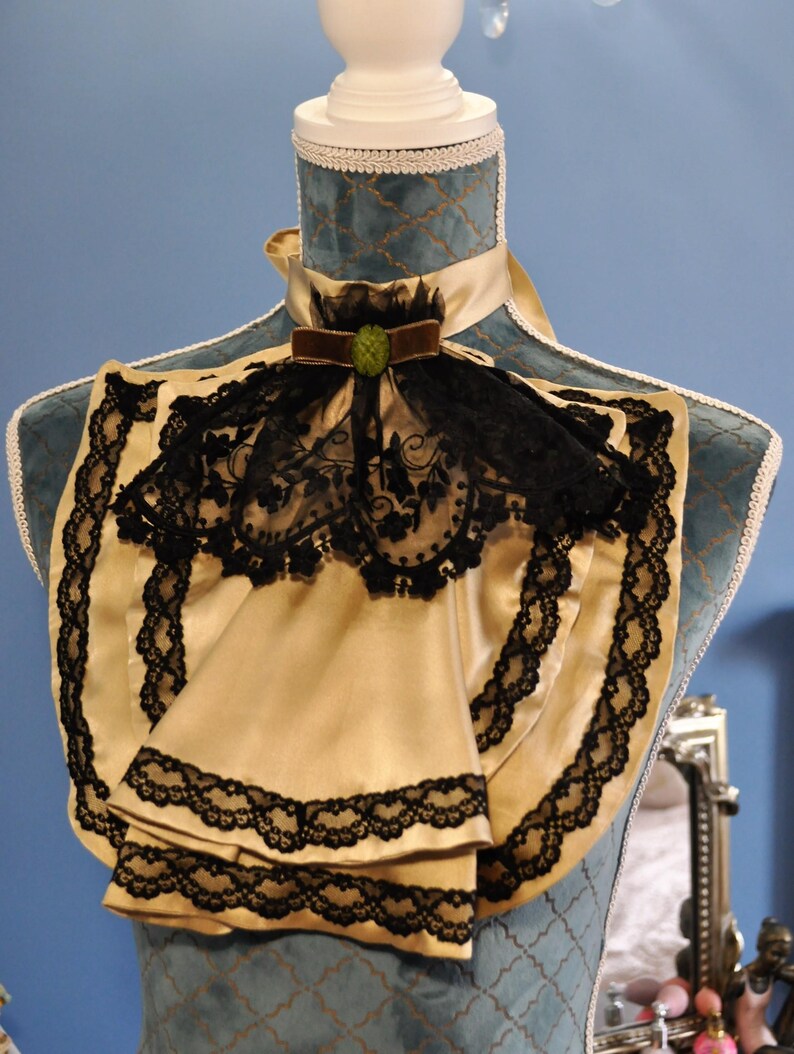 May include: A cream-colored satin ascot with black lace trim and a black lace overlay. The ascot features a brown velvet bow with a green jewel and is displayed on a blue mannequin.