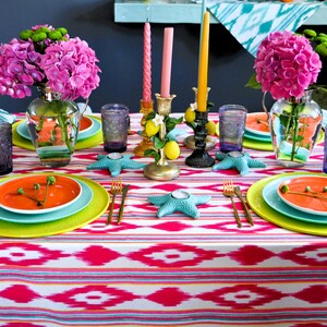 Peut inclure: Une table dress&eacute;e de mani&egrave;re color&eacute;e avec des hortensias roses, des citrons et des bougeoirs en forme d'&eacute;toile de mer turquoise. La table est recouverte d'une nappe ikat rose et blanche avec des assiettes vertes et orange.