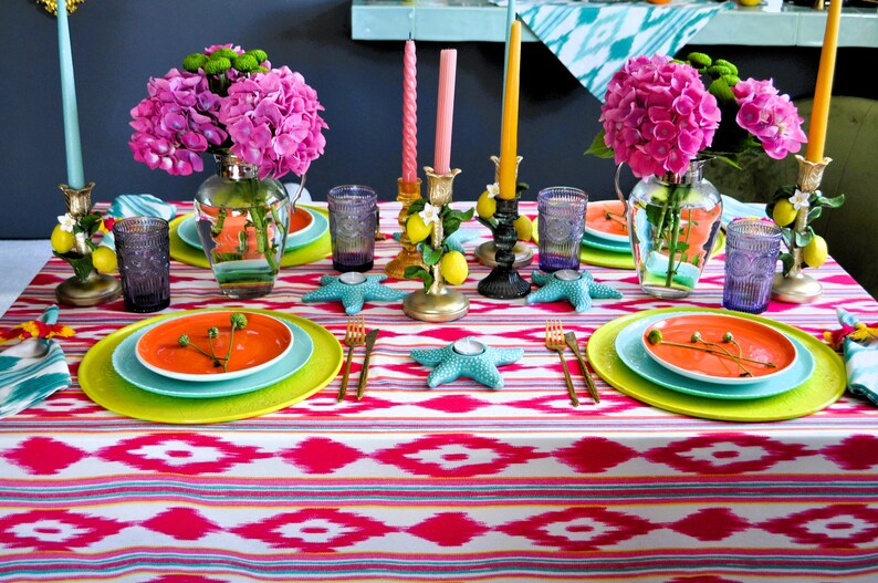 Peut inclure: Une table dress&eacute;e de mani&egrave;re color&eacute;e avec des hortensias roses, des citrons et des bougeoirs en forme d'&eacute;toile de mer turquoise. La table est recouverte d'une nappe ikat rose et blanche avec des assiettes vertes et orange.