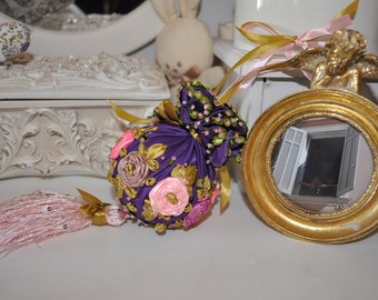 May include: A purple fabric ornament with pink and gold flowers and a pink tassel. The ornament is decorated with beads and sequins.