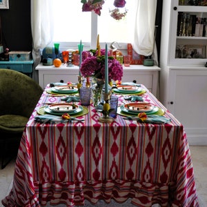 Peut inclure: Une nappe de table ray&eacute;e rose et blanche avec un bord festonn&eacute;, dress&eacute;e pour un d&icirc;ner. La table est d&eacute;cor&eacute;e d'assiettes vertes et orange, de bougies bleues et jaunes, et d'un bouquet de fleurs roses.