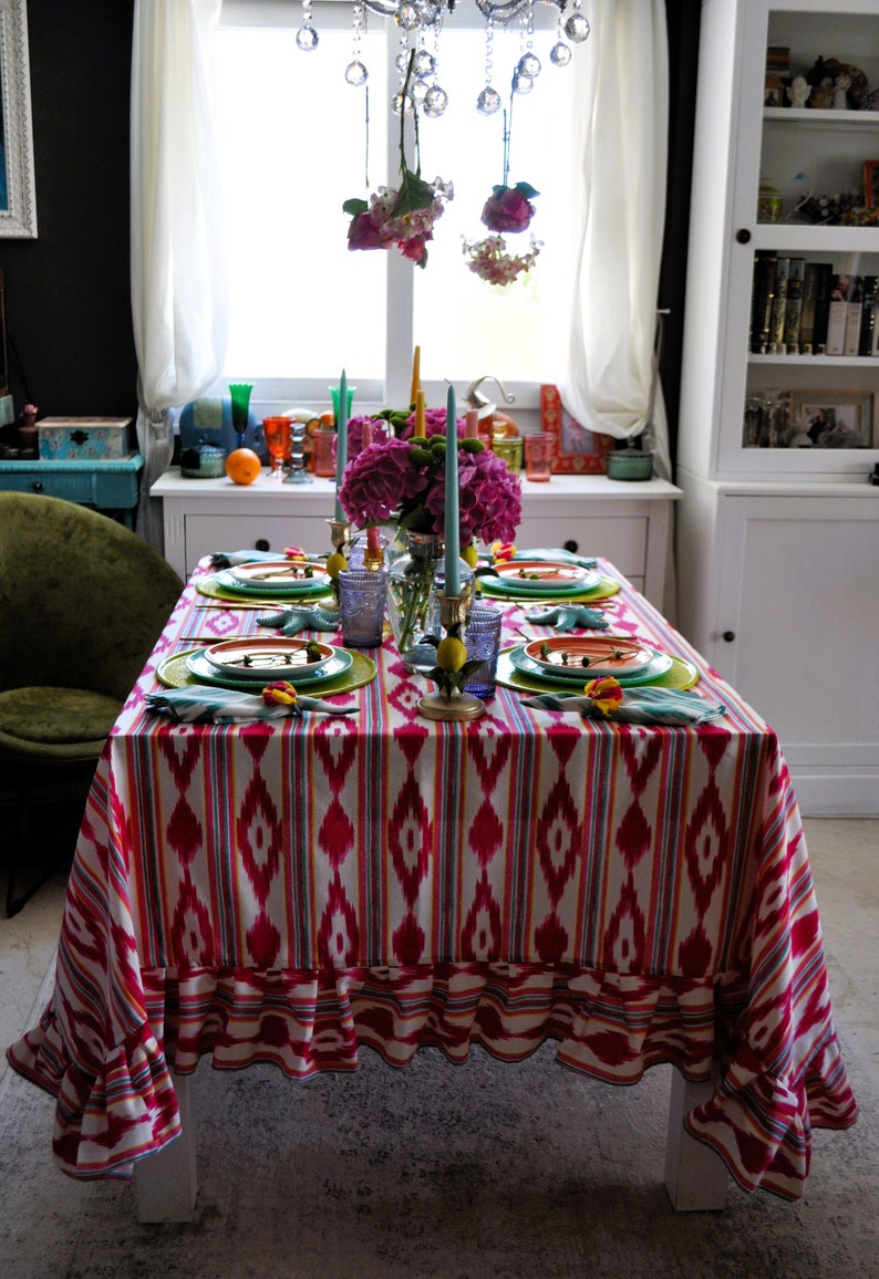 Peut inclure: Une nappe de table ray&eacute;e rose et blanche avec un bord festonn&eacute;, dress&eacute;e pour un d&icirc;ner. La table est d&eacute;cor&eacute;e d'assiettes vertes et orange, de bougies bleues et jaunes, et d'un bouquet de fleurs roses.
