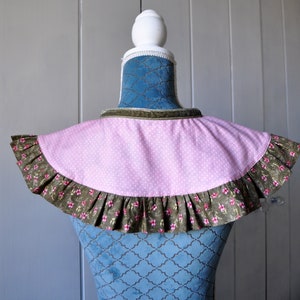 May include: A pink and white polka dot fabric collar with a brown floral ruffle trim. The collar is displayed on a blue velvet mannequin.