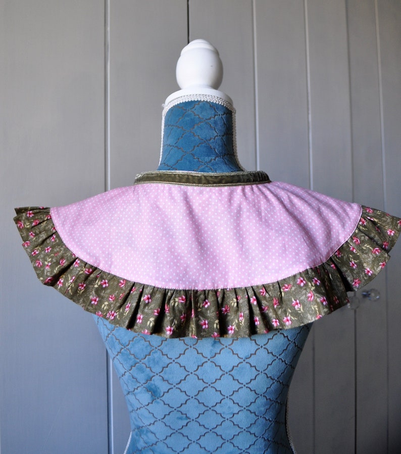 May include: A pink and white polka dot fabric collar with a brown floral ruffle trim. The collar is displayed on a blue velvet mannequin.