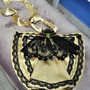May include: A gold satin apron with black lace trim and a black lace overlay. The apron has a gold satin tie and is decorated with a brown velvet bow and a green jewel.