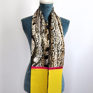 Peut inclure: Une longue écharpe fine avec un motif peau de serpent en brun, noir et blanc. L'écharpe a une bordure rose et jaune.